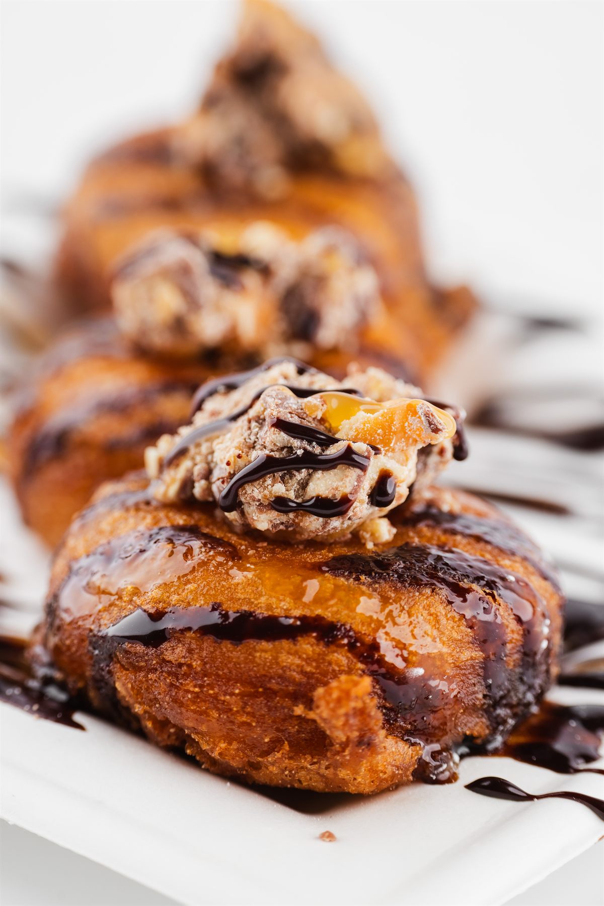 Three donuts on a plate under chocolate and caramel drizzle, topped with chopped candy.